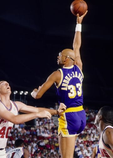Kareem Abdul-Jabbar executing his iconic skyhook shot during a game with the Los Angeles Lakers, wearing the purple and gold uniform, with defenders looking on.