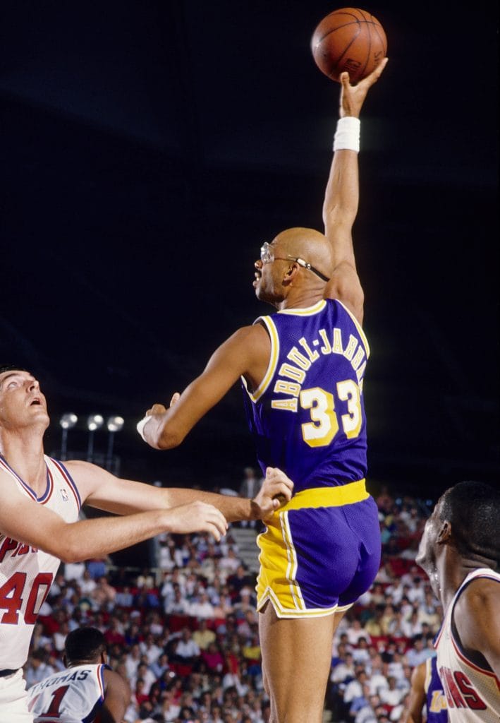 Kareem Abdul-Jabbar executing his iconic skyhook shot during a game with the Los Angeles Lakers, wearing the purple and gold uniform, with defenders looking on.