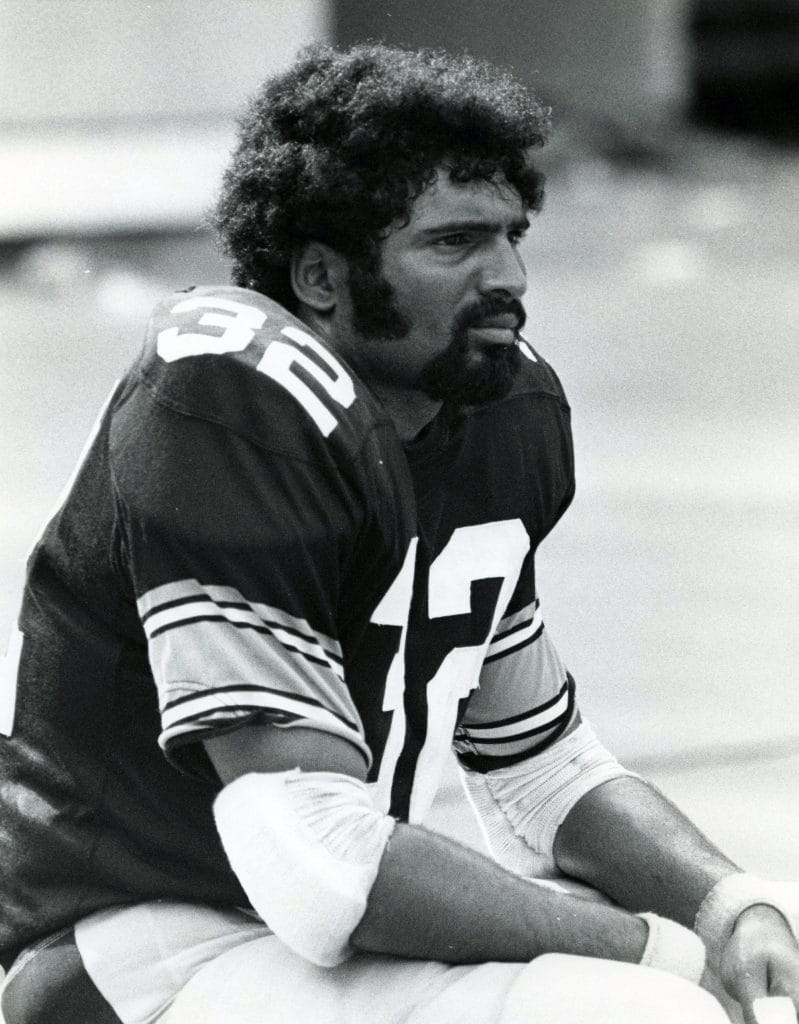Black-and-white image of Franco Harris of the Pittsburgh Steelers sitting on the sideline, appearing thoughtful during a game.