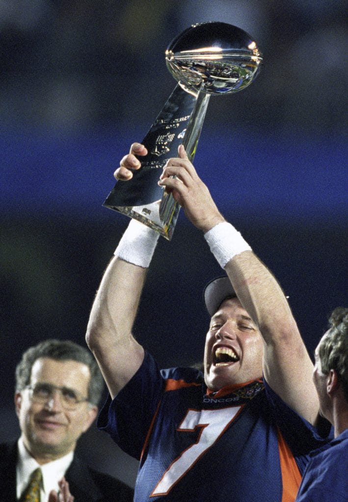 John Elway of the Denver Broncos holding the Lombardi Trophy in celebration after winning the Super Bowl, wearing jersey number 7.