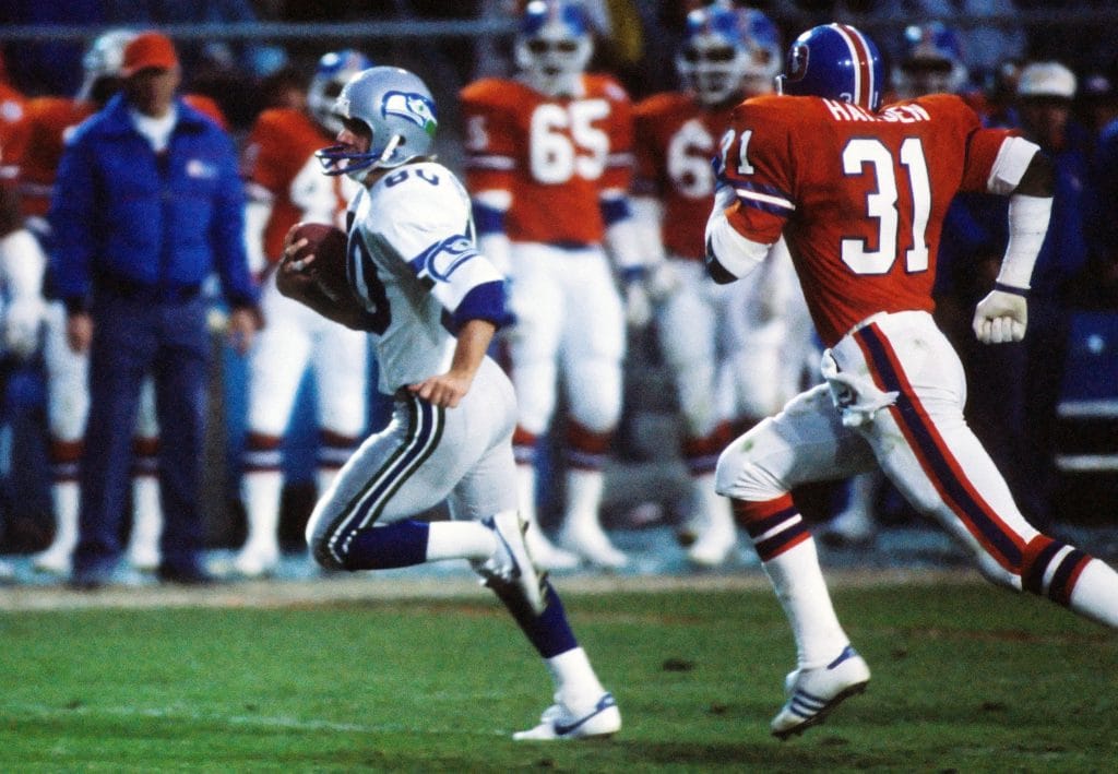 Seattle Seahawks player Steve Largent runs with the football while being pursued by a Denver Broncos defender during a game. One of the Top 10 Seattle Seahawks of All Time. 