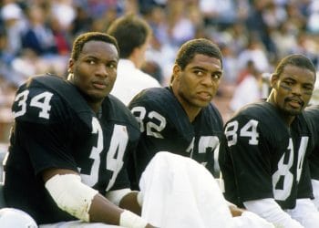 Bo Jackson (#34) and Marcus Allen (#32) seated on the Raiders' sideline in black uniforms during a game representing Top 10 Las Vegas Raiders of All Time.