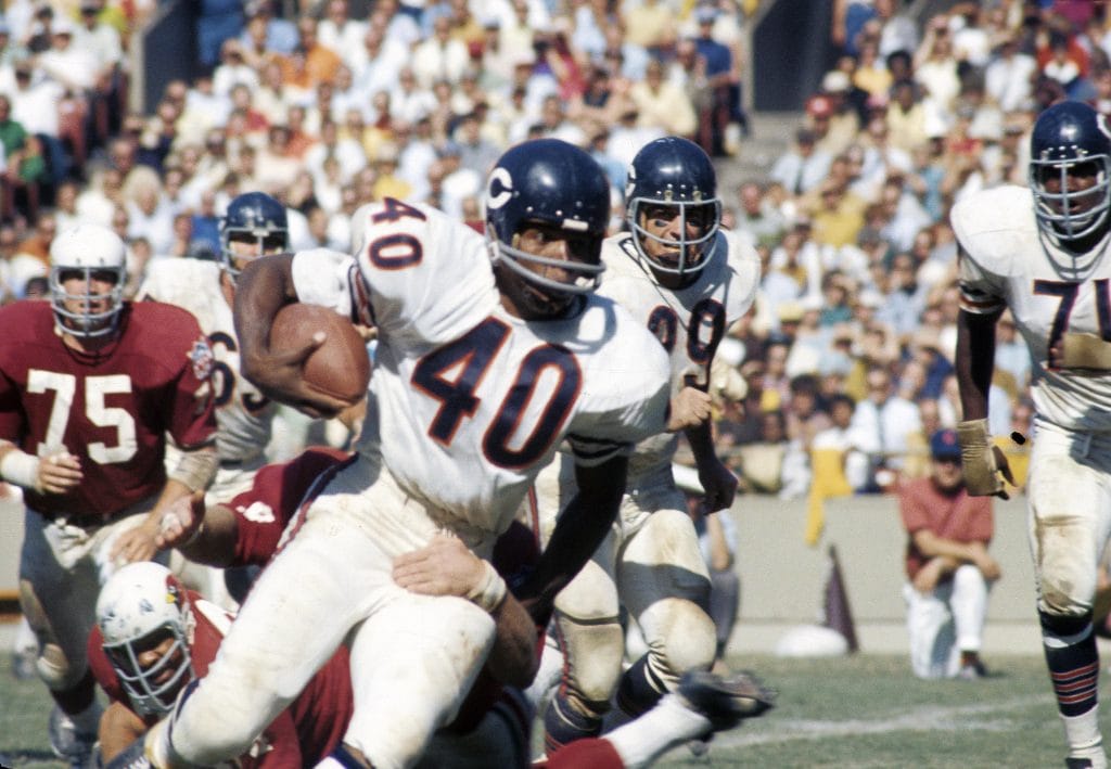 Gale Sayers in a Bears uniform, carrying the ball and evading defenders during an intense game.
