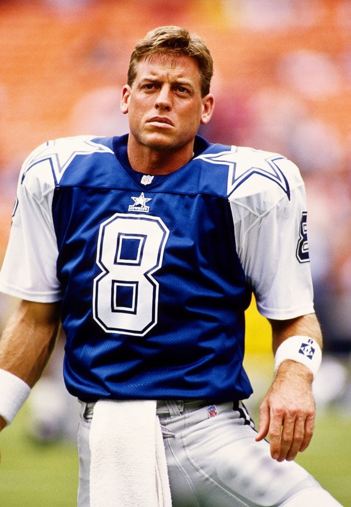 Dallas Cowboys quarterback Troy Aikman on the sidelines, focused before a game, wearing the iconic No. 8 jersey.