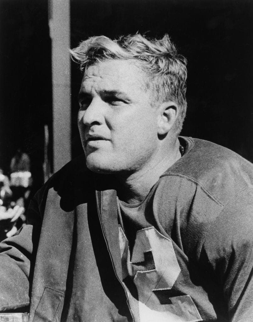 Bobby Layne seated on the sidelines in his football uniform, staring thoughtfully into the distance.