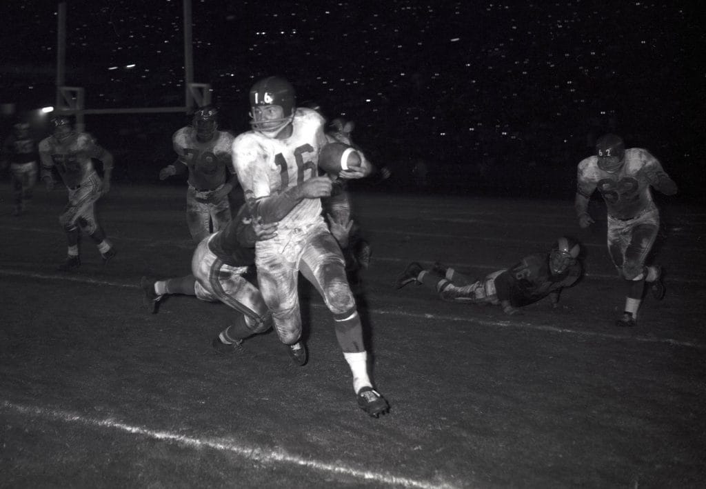 Frank Gifford wearing number 16 evades defenders during a classic NFL game under stadium lights.