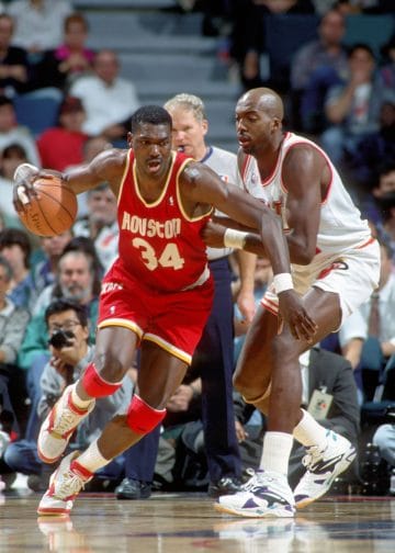 Hakeem Olajuwon aggressively driving to the basket against a defender during a Houston Rockets game showing why he is one of the Top 10 NBA Centers of All Time