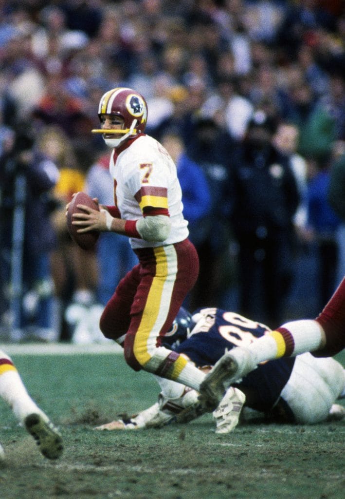 Joe Theismann of the Washington Redskins, wearing jersey number 7, evading a defender during an NFL game.