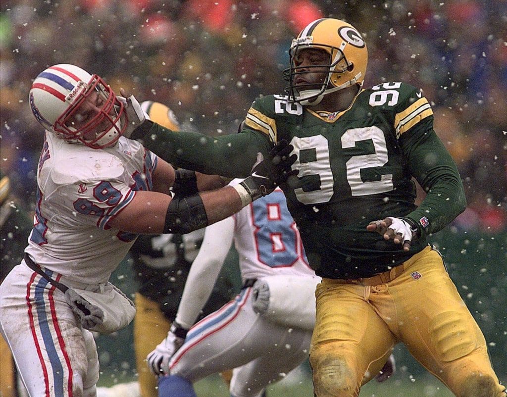 Reggie White, the Green Bay Packers' defensive legend, heads slaps an opposing lineman during a snowy game, displaying his signature power.