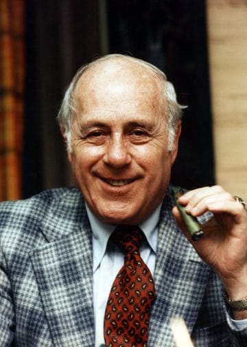 Red Auerbach, one of the best white coaches in NBA history, famous for his time with the Boston Celtics, holding a cigar as a symbol of victory.