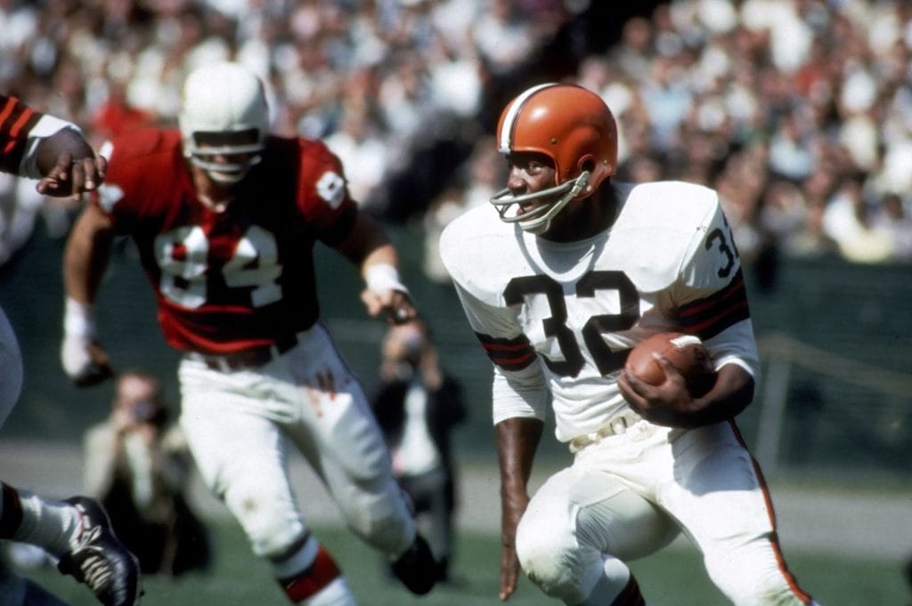 Jim Brown carrying the football during a game, showcasing his dominance as one of the Top 10 Running Backs Of All Time.