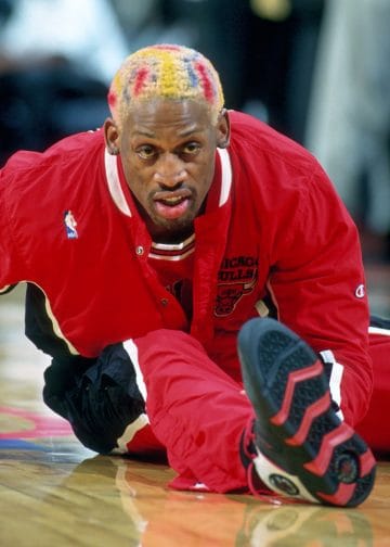 Close-up of Dennis Rodman stretching in a Chicago Bulls warmup with multicolored hair.
