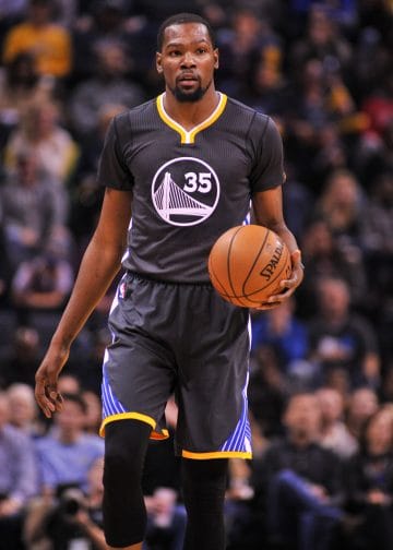 Kevin Durant bringing the ball up court in a black Golden State Warriors jersey. 