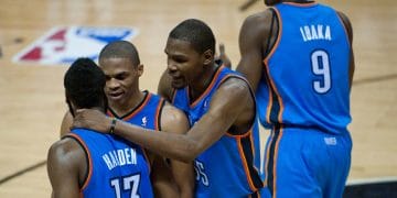 Kevin Durant, Russell Westbrook, and James Harden celebrating together on the court in Oklahoma City Thunder uniforms with Serge Ibaka in the background. 