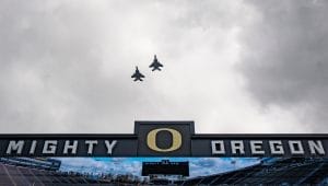Autzen Stadium flyover with Mighty Oregon scoreboard under cloudy skies.