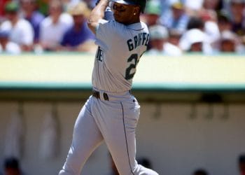 Black baseball players Ken Griffey Jr. finishing a powerful swing in a Seattle Mariners road uniform.