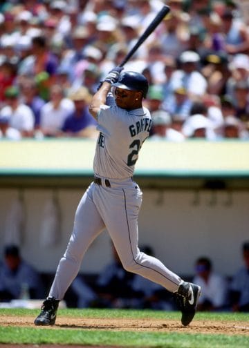 Black baseball players Ken Griffey Jr. finishing a powerful swing in a Seattle Mariners road uniform.