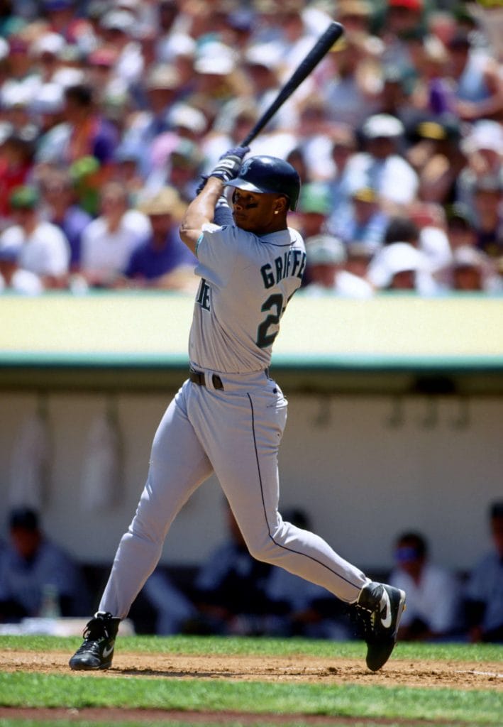 Black baseball players Ken Griffey Jr. finishing a powerful swing in a Seattle Mariners road uniform.