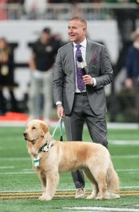 Kirk Herbstreit smiling on the College Football Playoff field with a golden retriever wearing credentials