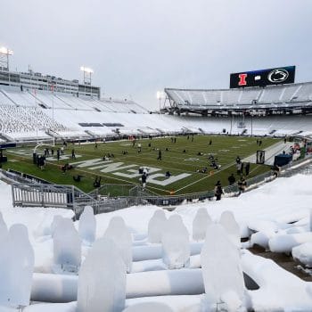 Penn State players warm up on a snowy field before a game against Illinois, with cardboard cutouts in the stands during a limited attendance season.