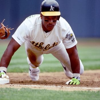 Black MLB legend Rickey Henderson leading off from first base in his Oakland Athletics uniform. 